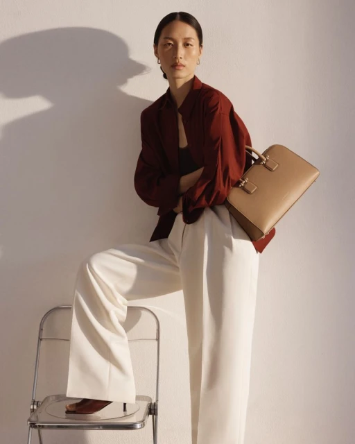 A sophisticated Asian woman poses indoors wearing a red blouse, white pants, and carrying a tan briefcase with a chair prop.