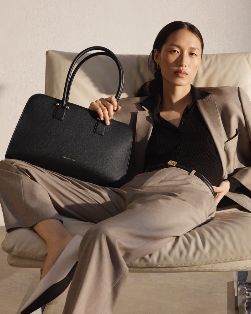 Woman in a suit sits holding a black leather tote bag indoors in a minimalist studio with natural light and neutral colors.