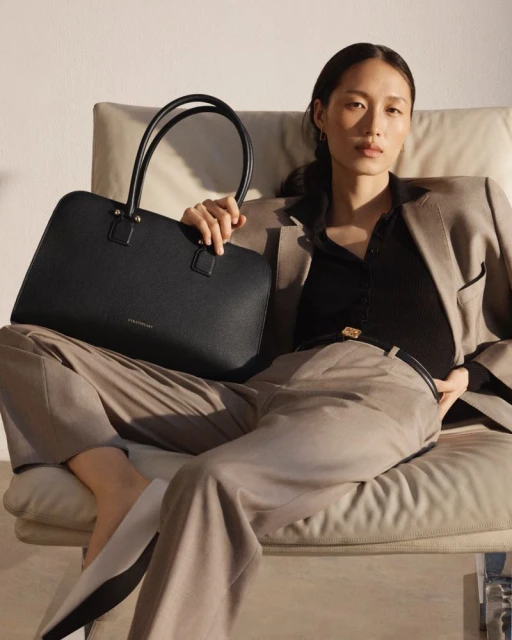 Woman in a suit sits holding a black leather tote bag indoors in a minimalist studio with natural light and neutral colors.