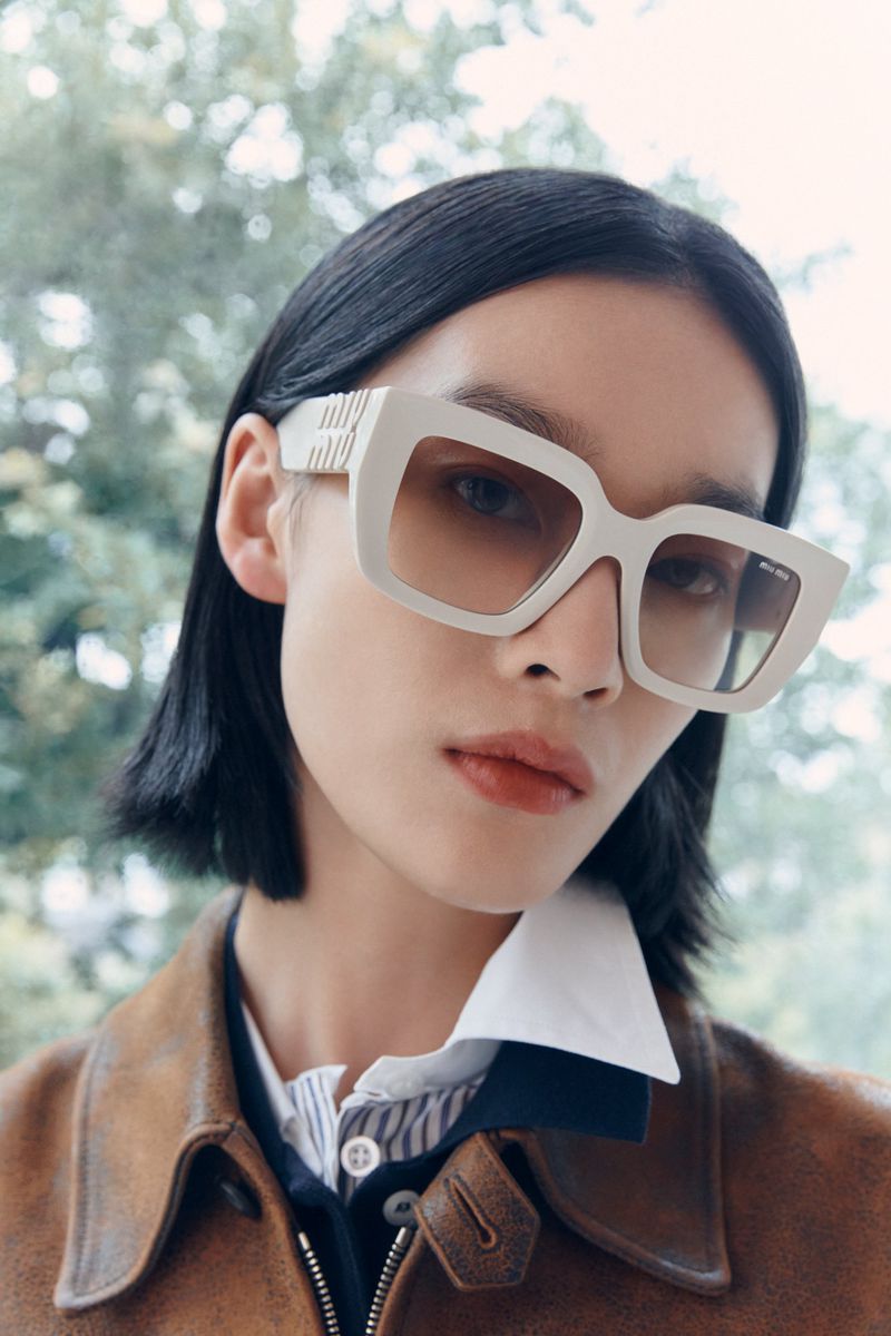 Fashionable portrait of a young adult woman with short dark hair wearing oversized white sunglasses and a brown leather jacket outdoors.