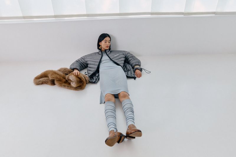 Asian female fashion model lying on a white surface with a gray puffer jacket, light blue dress, leg warmers, and brown shoes.