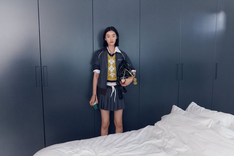 A young Asian woman stands in a pleated skirt and argyle sweater with a bag in an indoor bedroom setting against a blue closet door.