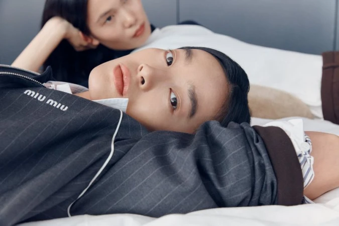 Close-up shot of two Asian models lying on a white bed. One is in focus, while the other is in the background, wearing a Miu Miu jacket.