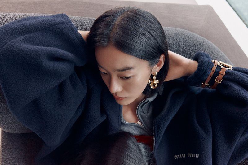 Eye-level shot of a woman in a navy fleece jacket with gold earrings and a brown watch, resting on a gray sofa.