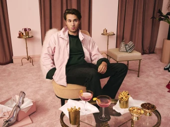 Man in pink jacket and dark trousers seated in an opulent pink room with cocktails and snacks on a glass table foreground