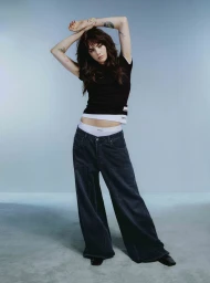 Model with long dark hair wearing a black t-shirt and wide-leg dark jeans standing against a plain grey studio background.