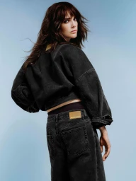 Model with long dark hair wearing a black denim jacket and matching jeans looking over her shoulder