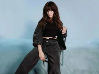 Woman with long dark hair and bangs seated on a dark metal chair wearing dark denim jacket, black top, and dark denim jeans