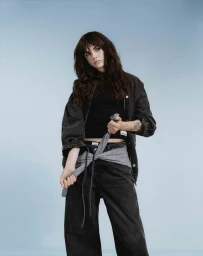 Woman in dark grey denim jacket and jeans holding a tied patterned shirt against a pale blue background