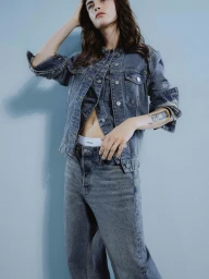 Model wearing a dark blue denim jacket and matching jeans posing against a plain blue background for Armedangels