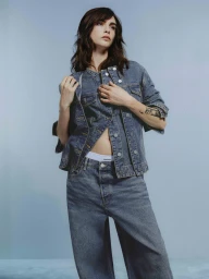 Woman in denim jacket and matching jeans posing against a light blue studio background for Armedangels