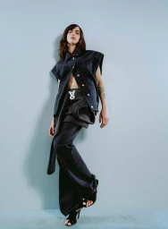 Model wearing a dark denim vest and black trousers leaning against a pale blue background for Armedangels