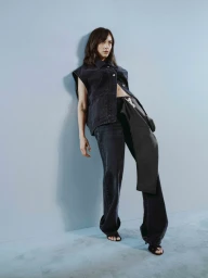 Woman in dark denim sleeveless vest and jeans leaning against a pale blue wall