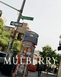 Grey leather handbag resting on a red street post covered in stickers beneath a Mulberry St street sign