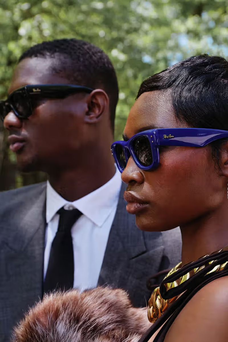 A fashionable couple poses outdoors: a man in a grey suit and sunglasses stands behind a woman in blue sunglasses and a gold necklace.