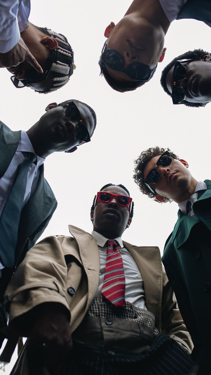Group of stylish adult men looking down at the camera, wearing formal attire, ties, sunglasses, and trench coats against white sky.