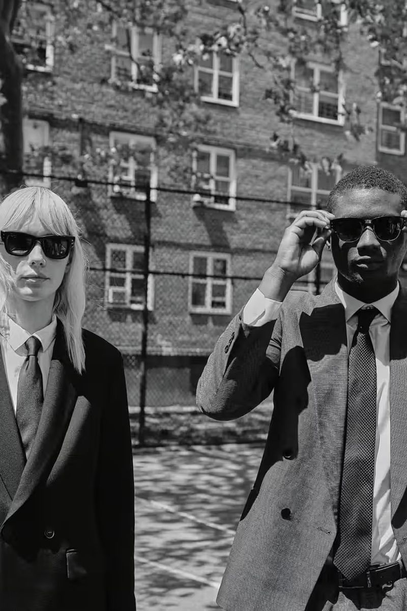 Black and white shot of a stylish adult man and woman wearing sunglasses and suits standing outdoors near an apartment building.