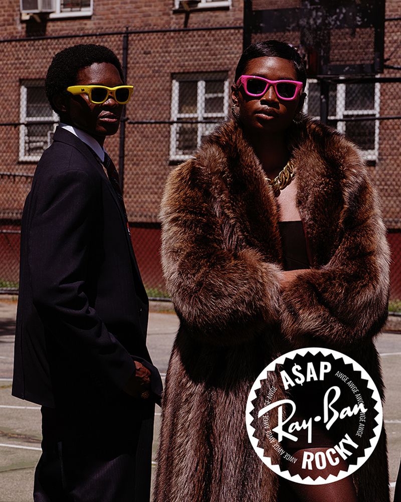 Two adult men pose outdoors against a brick wall in a fashion editorial. One man wears a fur coat and the other a dark suit.