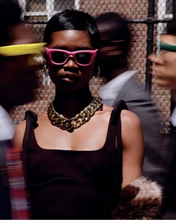 Stylish woman in brown dress and pink sunglasses stands out in a blurry urban street scene, accessorized with a chunky gold necklace.