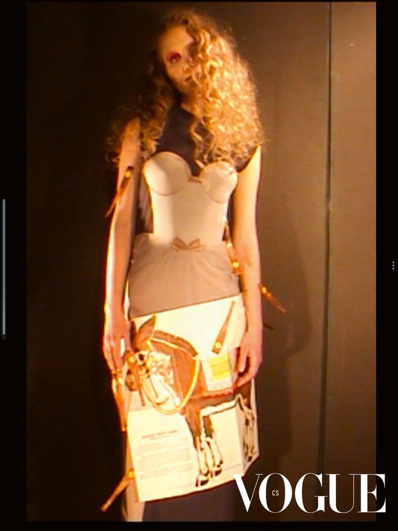 A fair-skinned adult woman stands with curly brown hair, wearing a conceptual fashion garment in front of a solid black background.