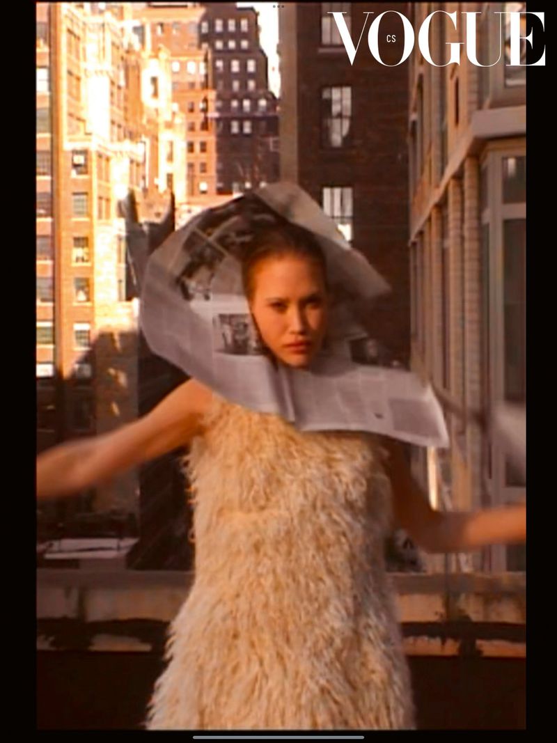 A woman in a shaggy dress and newspaper collar stands on a rooftop with city buildings in the background, Vogue logo in the top right.
