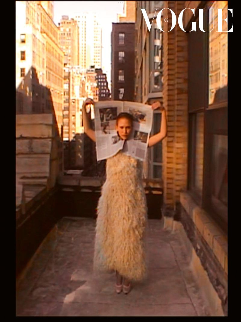 Fashion shoot featuring a young woman in a furry dress holding a newspaper on a city rooftop with tall buildings in the background.