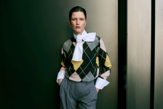 Model wearing a patterned argyle sweater and grey trousers posing against a dark green wall for Harrods.
