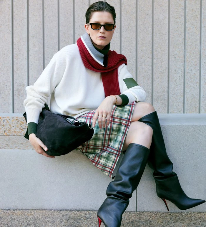 Model wearing dark sunglasses, white sweater, red scarf, plaid skirt, and black boots holding a black handbag.