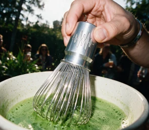 Close-up of a person using a metallic matcha whisk with soundwave engraving inside a ceramic bowl filled with bright green matcha.