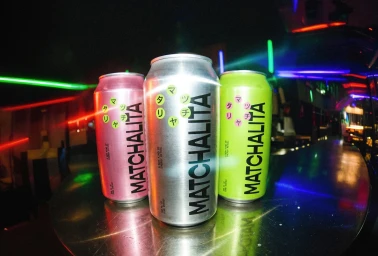 Three beverage cans branded Matchalita in pink, silver, and lime green on a metal bar counter with glowing neon lights in the background.