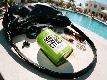 Green tea canister next to an open black leather bag by a swimming pool with sunglasses, lipstick, and earbuds