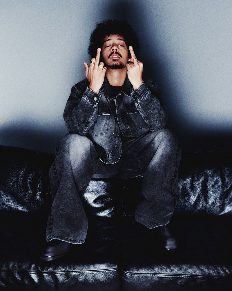 Person with afro hairstyle sits on black leather couch wearing denim jacket and matching jeans, with two middle fingers extended.