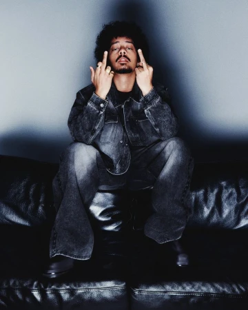 Person with afro hairstyle sits on black leather couch wearing denim jacket and matching jeans, with two middle fingers extended.