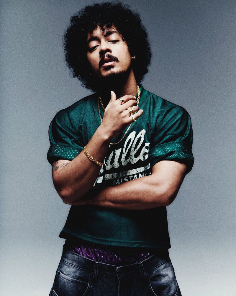 Portrait of a young adult man with dark curly hair wearing a green sports jersey, gold jewelry, and blue jeans against a gray background.