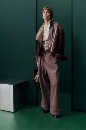 Model in a brown leather jacket and wide-leg trousers standing in a green studio set with a metallic cube.