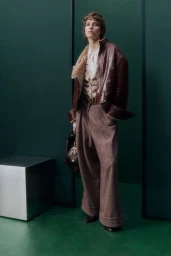 Model in a brown leather jacket and wide-leg trousers standing in a green studio set with a metallic cube.