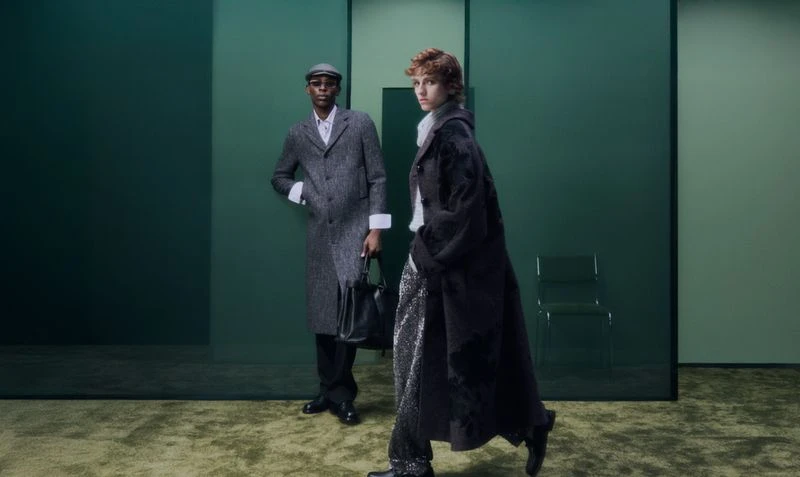 Two models wearing dark winter coats standing against a green-toned studio background with a green chair