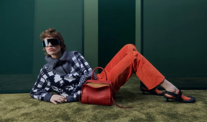 Model wearing dark visor sunglasses and black and white patterned sweater posing with a red handbag
