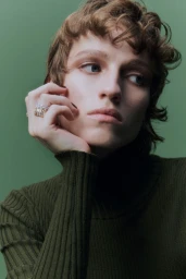 Model wearing a dark green ribbed turtleneck sweater resting chin in hand against a solid green background
