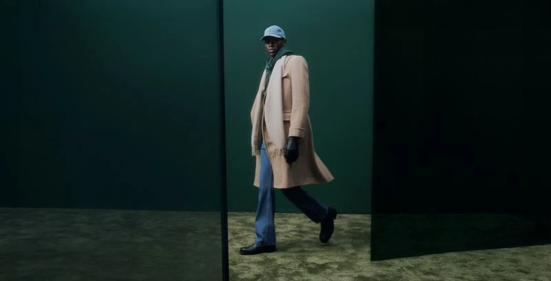 Model Adamu Bulus wearing a tan coat, cap, and blue jeans against a dark studio background for Harrods.