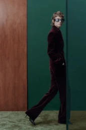 Model wearing dark sunglasses and a burgundy velvet suit walking against a green and wood-paneled wall