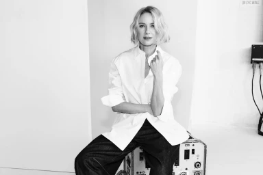 Naomi Watts seated on equipment cases wearing a white button-down shirt and dark trousers against a white backdrop.