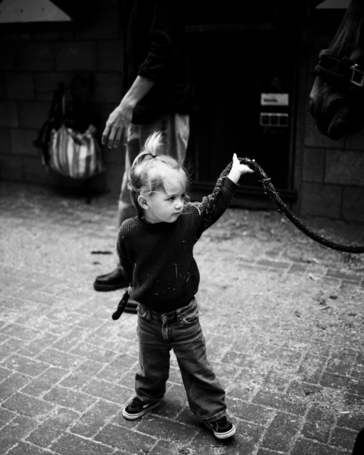 Monochrome shot of a toddler holding a rope, with an adult and horse visible in the background, taken on a brick surface.