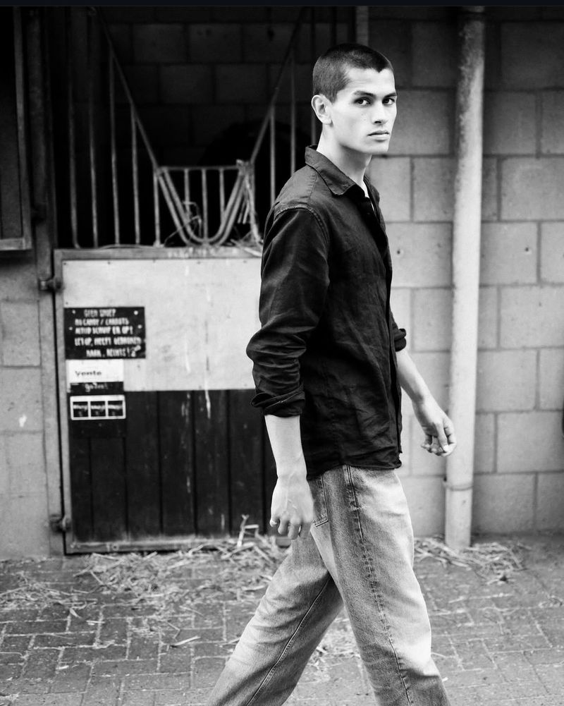 Monochrome portrait of a young adult male walking outdoors in an open shirt and jeans, with a building and doorway in the background.