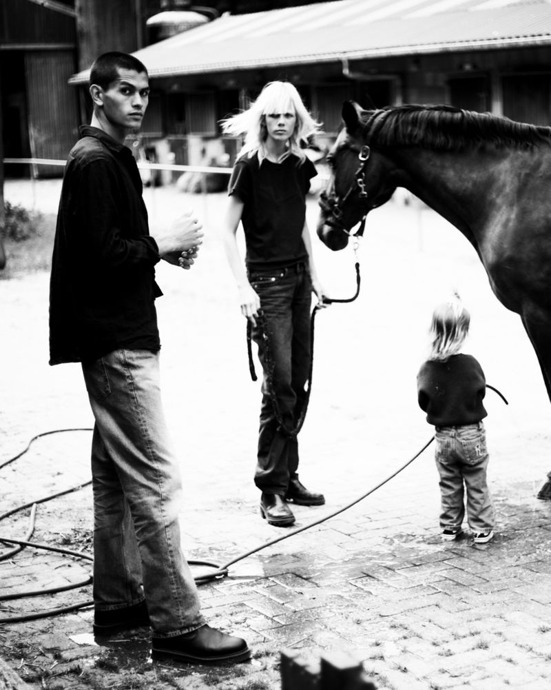 Monochrome outdoor portrait of two adult humans and a child standing near a horse, with brick ground and a barn in the background.