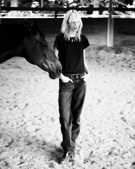 Black and white full shot of a female with blonde hair wearing jeans and boots, standing next to a horse in an outdoor paddock.