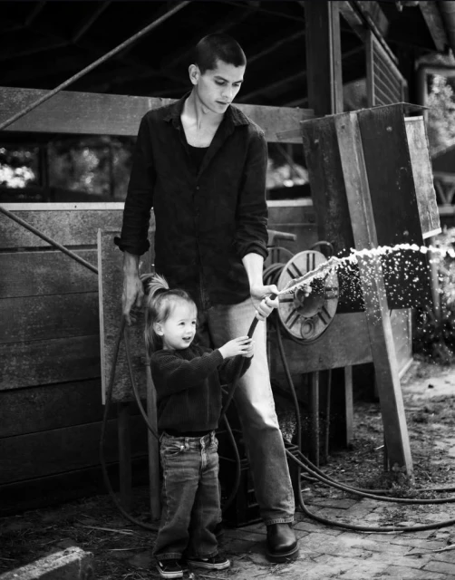 Young man and a toddler girl holding a garden hose together, spraying water outdoors. Black and white lifestyle photography.