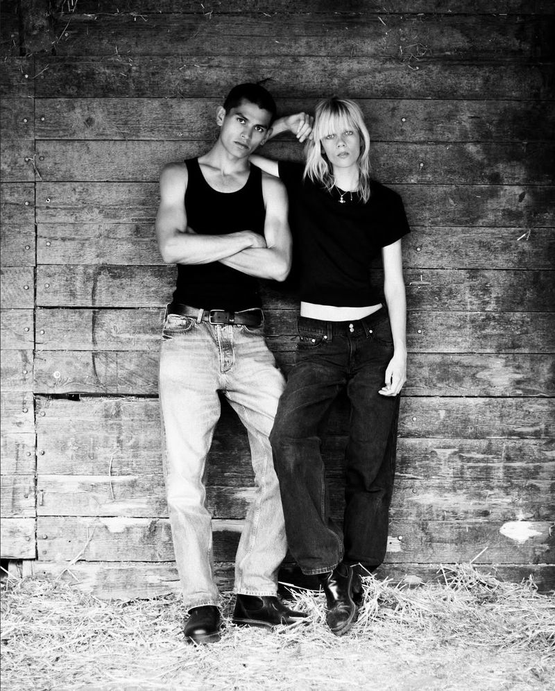 Monochrome portrait of a young couple leaning against a weathered wooden wall outdoors, evoking a vintage or documentary feel.