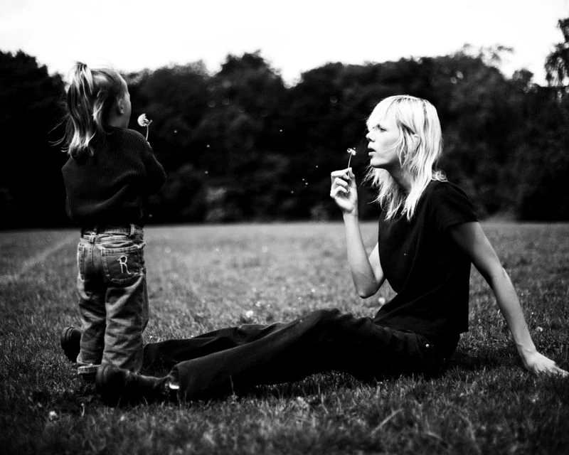 A child and an adult woman are sitting in a grassy field blowing dandelions in a black and white outdoor scene with trees in the background.