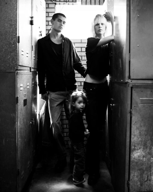 Black and white portrait of an adult male, adult female, and child standing in a hallway with metal lockers, looking at the camera.